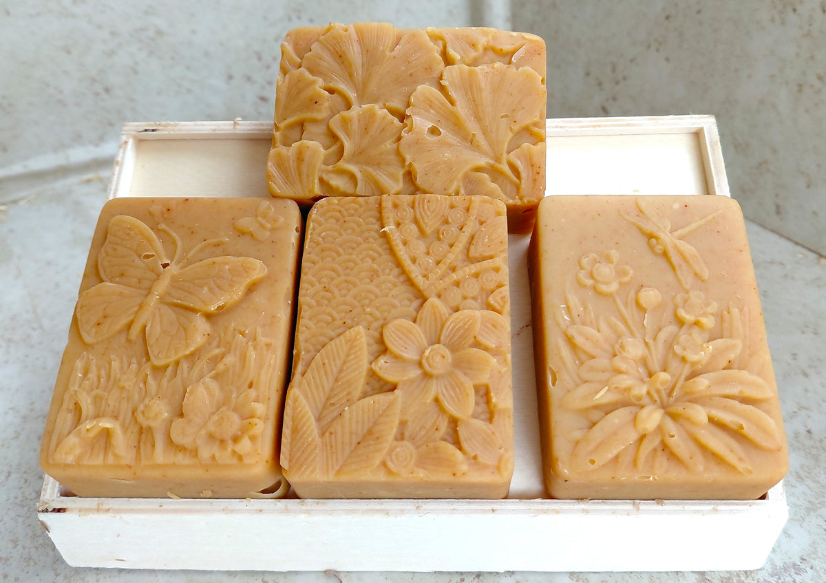 Four rectangular bars of natural tan soap with embossed floral and butterfly designs, arranged in a white wooden tray on a light stone surface.