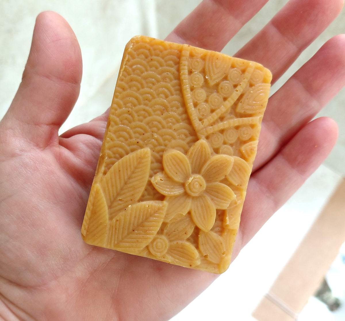 A hand holding a rectangular, golden-yellow soap bar with embossed floral and wave patterns.