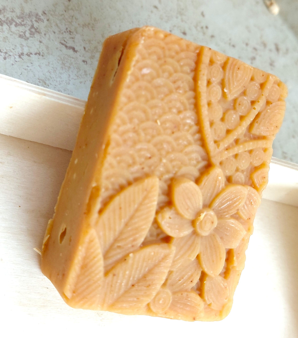 A rectangular bar of handmade soap in a warm tan or light brown color, featuring embossed floral and leaf patterns on one side, resting on a white surface.