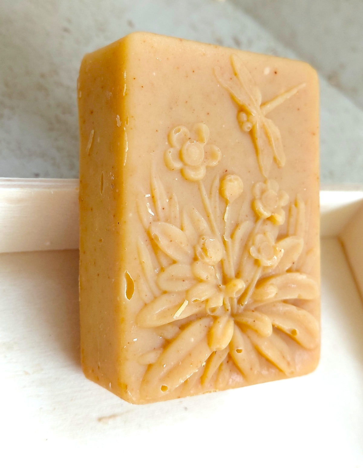 A rectangular bar of handmade soap in a warm tan or light brown color, embossed with floral and dragonfly designs, standing upright on a white surface.