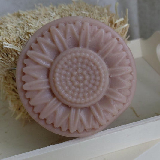 Pink sunflower-shaped soap bar with a textured surface, placed on a white stand against a neutral background.