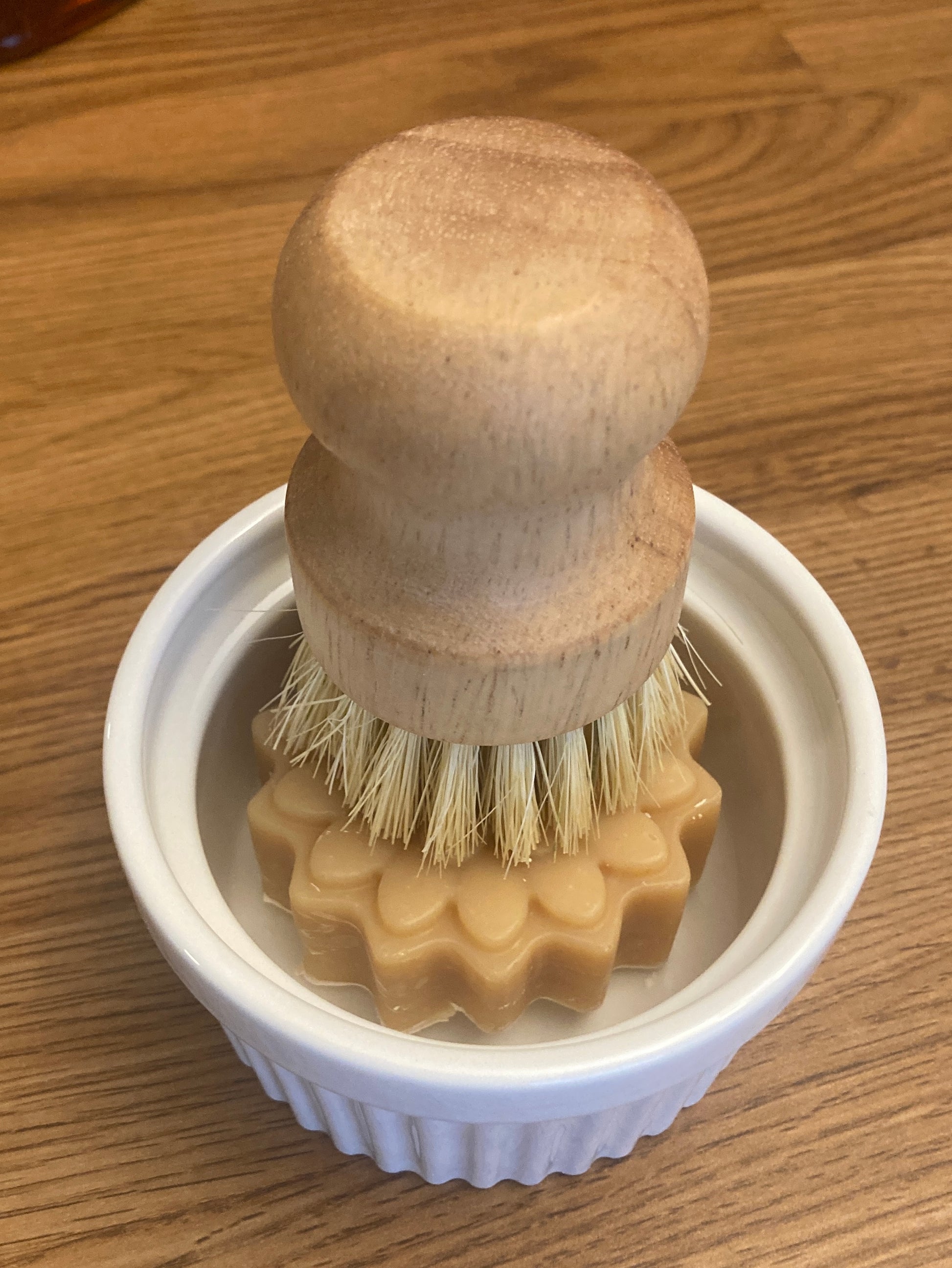 Wooden brush with bristles in a white ramekin with a beige solid dish soap bar on a wooden surface